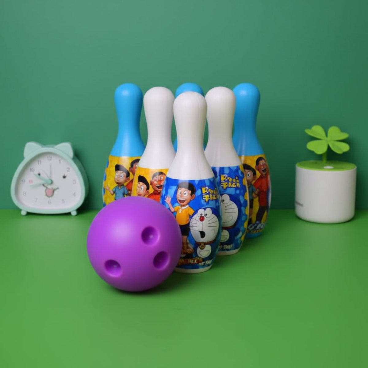 A purple bowling ball and a set of colorful toy bowling pins with cartoon graphics, displayed on a green surface with a small clock and plant in the background.